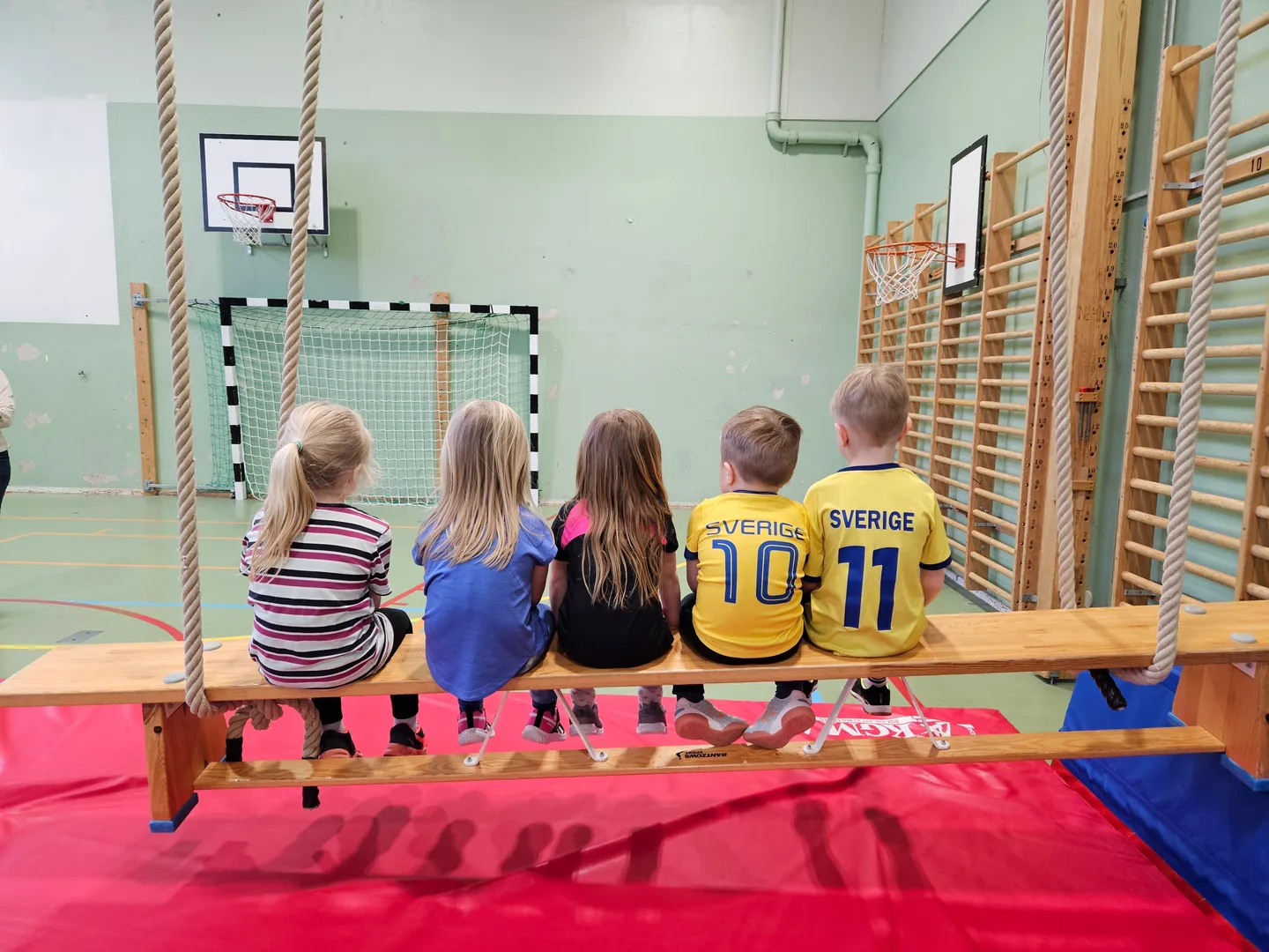 Fem barn sitter på en bänk i gympasalen på Högoms skola inför barngympan